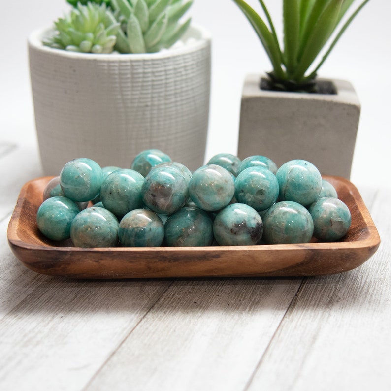May include: A wooden bowl filled with polished, teal-colored spheres. The spheres have a marbled appearance with darker spots. Two small potted plants are in the background. The bowl is made of a light brown wood.