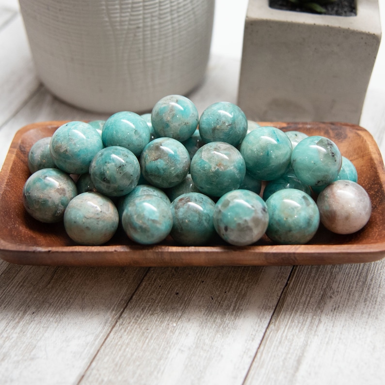May include: A wooden bowl filled with polished, spherical gemstones. The stones are primarily turquoise with mottled gray and brown inclusions. The bowl sits on a light-colored wooden surface, with a white and gray background.