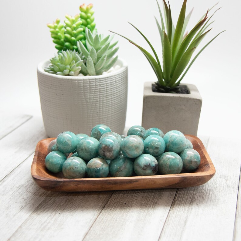 May include: A wooden tray filled with polished, round, turquoise-colored gemstones. The gemstones have a mottled appearance with darker spots. Two small potted succulents are in the background.