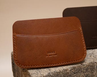May include: A brown leather cardholder with a curved top pocket and stitched edges. The cardholder has the text "BRIKS BY MATTONI" embossed on the front. Another darker brown cardholder is visible in the background.