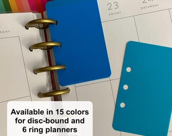 May include: A royal blue rectangular disc-bound planner insert with a notched edge and a separate cyan rectangular planner insert featuring three circular holes along the left side, both resting on a white planner page with grid lines and text.