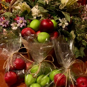 May include: A basket filled with red and green apples is surrounded by a floral arrangement. The apples are packaged in clear plastic bags tied with twine.