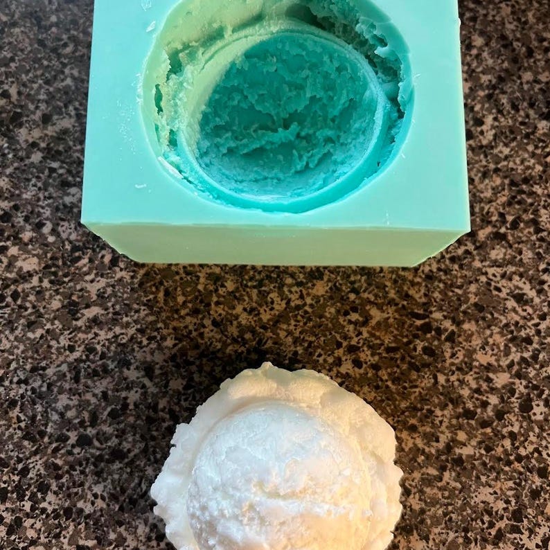 K&ouml;nnte beinhalten: Eine wei&szlig;e Silikonform zum Herstellen von Eiscreme oder anderen Desserts. Die Form ist halbrund und hat eine glatte, strukturierte Oberfl&auml;che. Die Form steht auf einer dunkelbraunen Arbeitsplatte.