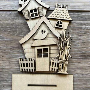 May include: A wooden Halloween-themed house decoration. The house features a slanted roof, windows, a door, and a ghost cutout. A small fence and bundle of corn stalks are also included. The base has a slot for display.