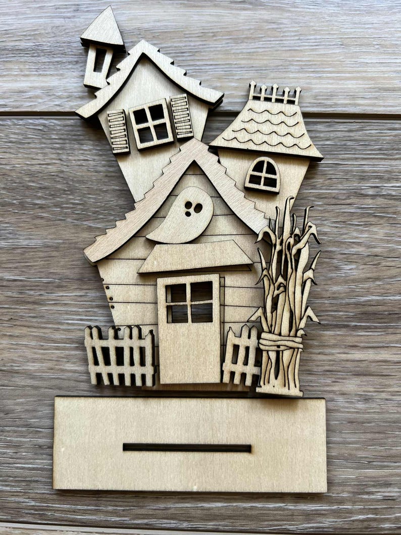 May include: A wooden Halloween-themed house decoration. The house features a slanted roof, windows, a door, and a ghost cutout. A small fence and bundle of corn stalks are also included. The base has a slot for display.