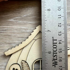 May include: A wooden Halloween-themed house with a ghost cutout and window. The house is approximately 5 inches tall, as measured by a Westcott ruler. The house is made of light-colored wood.
