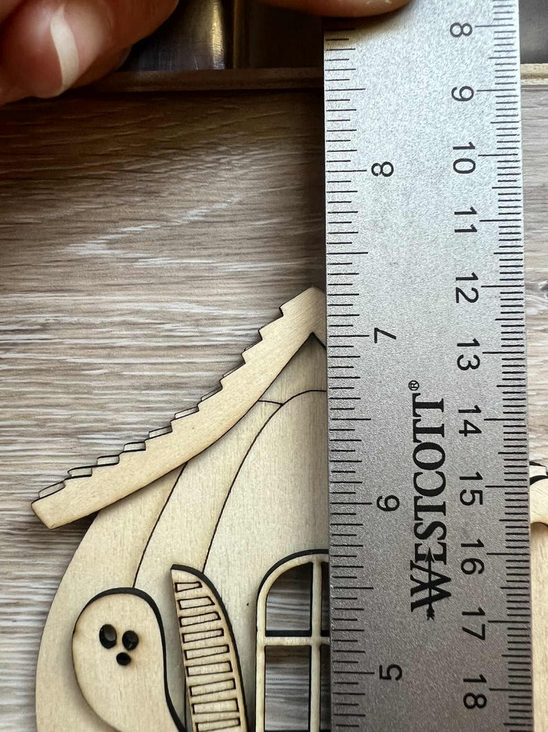 May include: A wooden Halloween-themed house with a ghost cutout and window. The house is approximately 5 inches tall, as measured by a Westcott ruler. The house is made of light-colored wood.