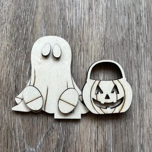 May include: Wooden Halloween decorations featuring a ghost and a jack-o'-lantern basket. The ghost has oval eyes and round feet. The jack-o'-lantern has a carved face and a handle. Both are made of light-colored wood.