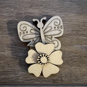 May include: A wooden cutout of a butterfly perched on a flower. The butterfly is detailed with swirls and dots. The flower has five petals and a center circle.