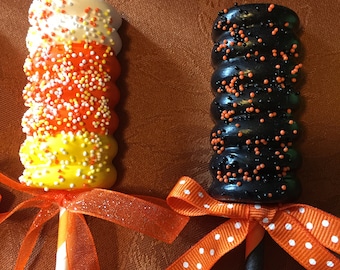Puede incluir: Dos cake pops de Halloween en palitos con pajitas de papel rayadas de naranja y blanco. El cake pop de la izquierda está cubierto de chispas blancas, naranjas y amarillas. El cake pop de la derecha está cubierto de chispas negras y naranjas. Ambos cake pops tienen cintas de lunares naranja y blanco atadas alrededor de las pajitas.