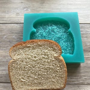 May include: A teal silicone mold shaped like a slice of bread. A slice of white bread is next to the mold.