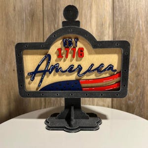 May include: A wooden sign with the word "America" in blue script, the text "EST 1776" in red, and a stylized American flag design. The sign has a dark gray frame and base, and is displayed on a white surface.