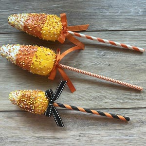 May include: Three candy corn-shaped treats on sticks, decorated with orange, yellow, and white sprinkles. Each treat has a ribbon and a decorative straw. The straws have orange and white, or black and orange stripes.