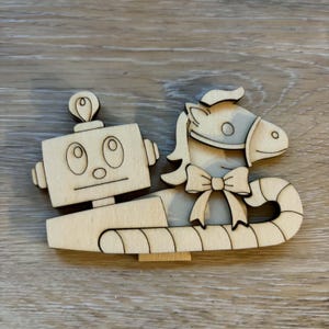 May include: A light-colored wooden cutout featuring a rectangular robot head with a small antenna on top, positioned next to a horse head adorned with a bow tie, all resting on a curved, segmented base that resembles a candy cane shape.