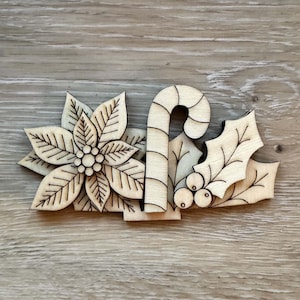 May include: A laser-cut wooden ornament featuring a poinsettia flower on the left, a candy cane in the center, and holly leaves with berries on the right. The design has engraved outlines on light-colored wood with a natural grain finish.