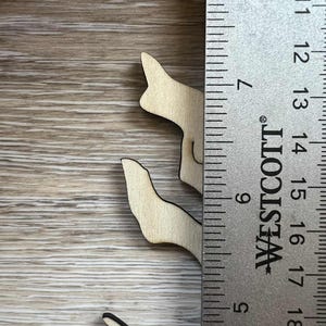 May include: Wooden cutouts of a stylized cat, approximately 3 inches tall, with a black outline. The cat cutouts are placed on a light brown wooden surface next to a ruler.