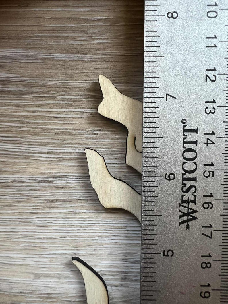 May include: Wooden cutouts of a stylized cat, approximately 3 inches tall, with a black outline. The cat cutouts are placed on a light brown wooden surface next to a ruler.
