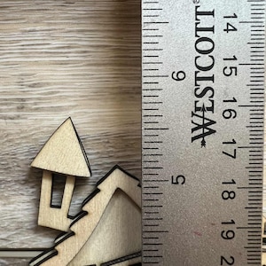 May include: A wooden house-shaped cutout with black trim, next to a ruler. The house has a chimney and windows. The ruler is marked with inches, showing the house's approximate height.