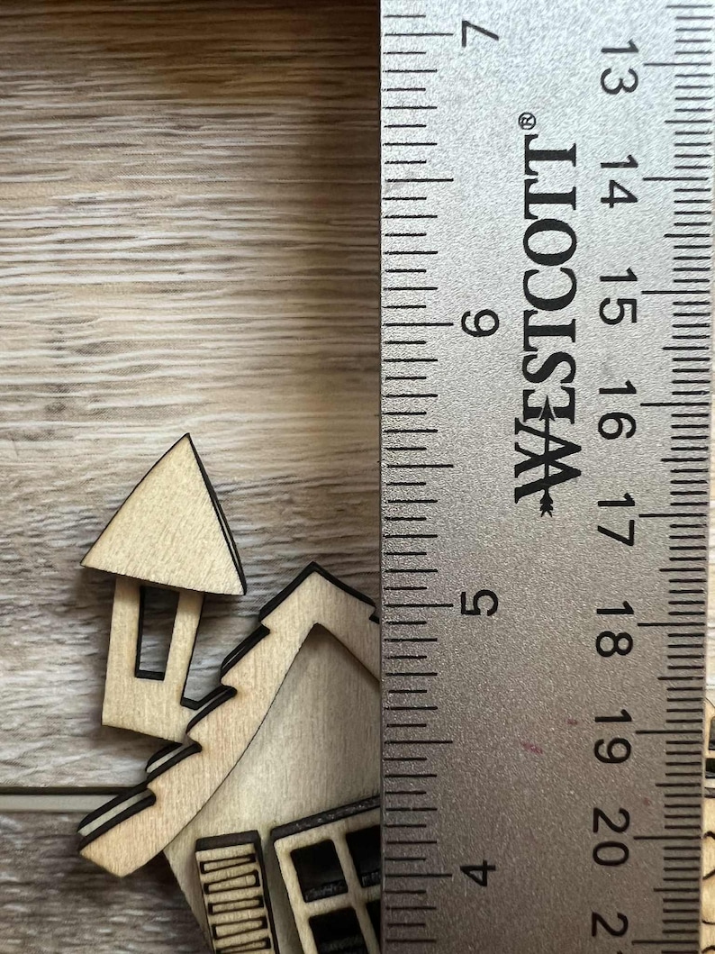 May include: A wooden house-shaped cutout with black trim, next to a ruler. The house has a chimney and windows. The ruler is marked with inches, showing the house's approximate height.