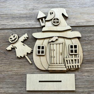 May include: A wooden Halloween decoration featuring a pumpkin-shaped house with a witch hat roof, windows, and a door. A scarecrow and a base are included. The decoration is made of unfinished wood.