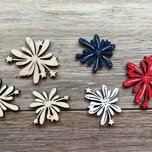 May include: Wooden firework-shaped cutouts in various colors. The cutouts include natural wood, navy blue with white accents, red with white accents, and white with white accents. Each firework has star accents.