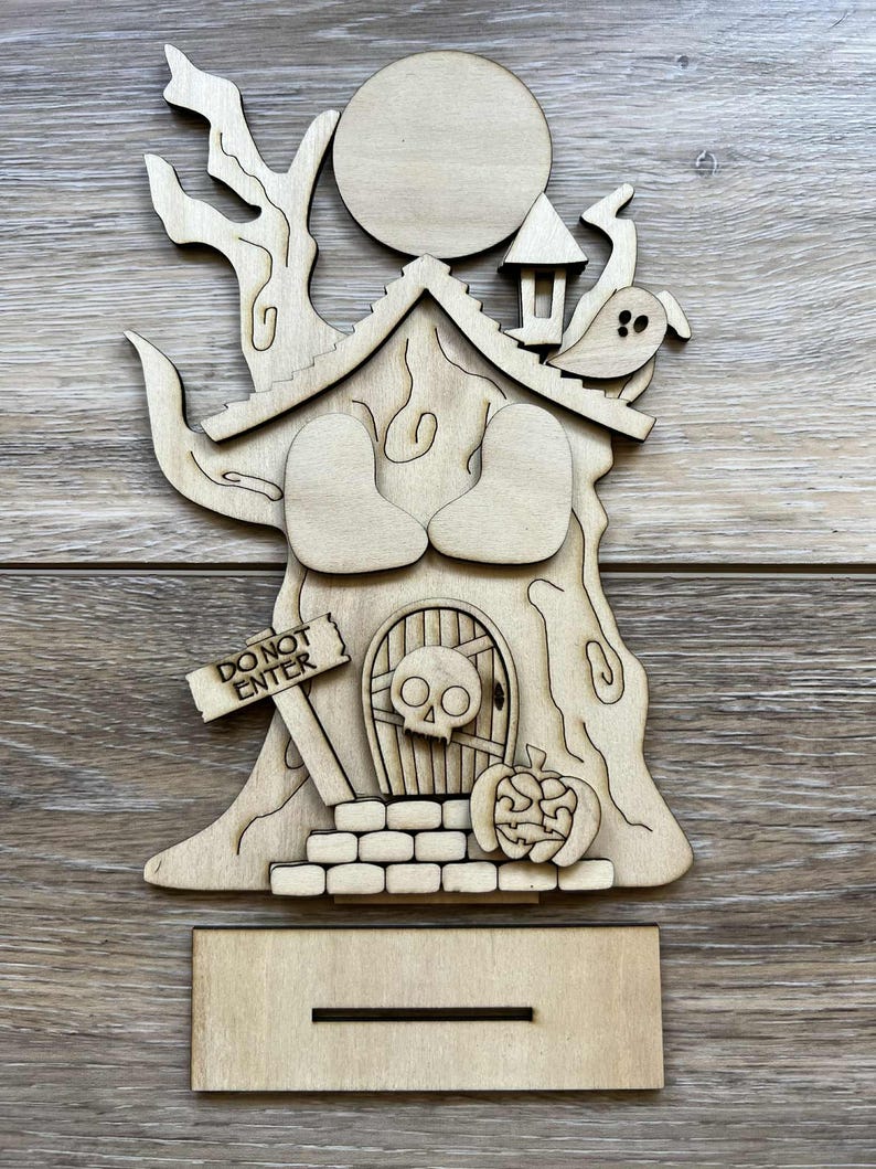 May include: A wooden Halloween-themed decoration featuring a tree with a house, a full moon, and a ghost. The house has a skull door and a pumpkin. The sign reads "DO NOT ENTER". Includes a stand.