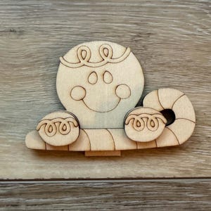 May include: A light-colored wooden cutout of a gingerbread person head with a smiling face, featuring a curved line representing hair at the top and two small circular hands resting on a horizontal candy cane-shaped base with striped details.