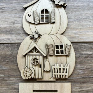 May include: A wooden Halloween-themed house decoration. The house is shaped like two stacked pumpkins, with a ghost, spider, and bat. Features include a door, windows, shutters, a chimney, and a small fence. The decoration is made of unfinished wood.