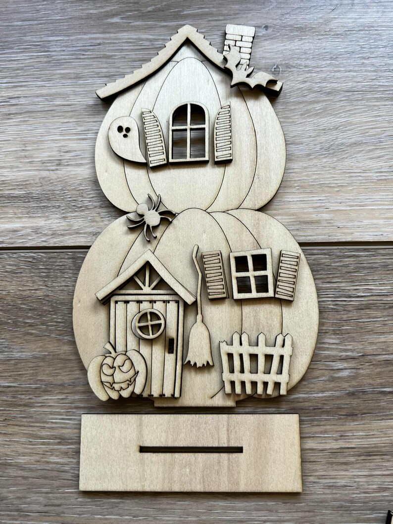 May include: A wooden Halloween-themed house decoration. The house is shaped like two stacked pumpkins, with a ghost, spider, and bat. Features include a door, windows, shutters, a chimney, and a small fence. The decoration is made of unfinished wood.