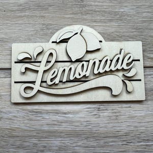 May include: Wooden sign with the word "Lemonade" in a cursive font. The sign features a lemon and leaves design above the word and black horizontal lines. The sign is made of light-colored wood.