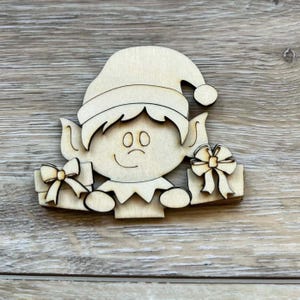 May include: A wooden Christmas elf cutout with a hat, ears, and a smiling face. The elf holds two gift boxes with bow accents. The cutout is made of light-colored wood and is suitable for holiday crafts.