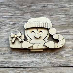 May include: Wooden penguin Christmas ornament holding a gift box with a bow and a candy cane. The penguin wears a Santa hat. The ornament is made of light-colored wood and has a cheerful expression.