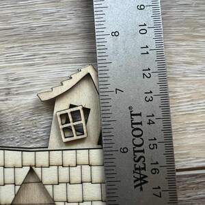 May include: A wooden miniature house with a detailed roof, window, and chimney. A metal ruler is placed next to the house, showing a height of approximately 2.5 inches. The house is made of light-colored wood.