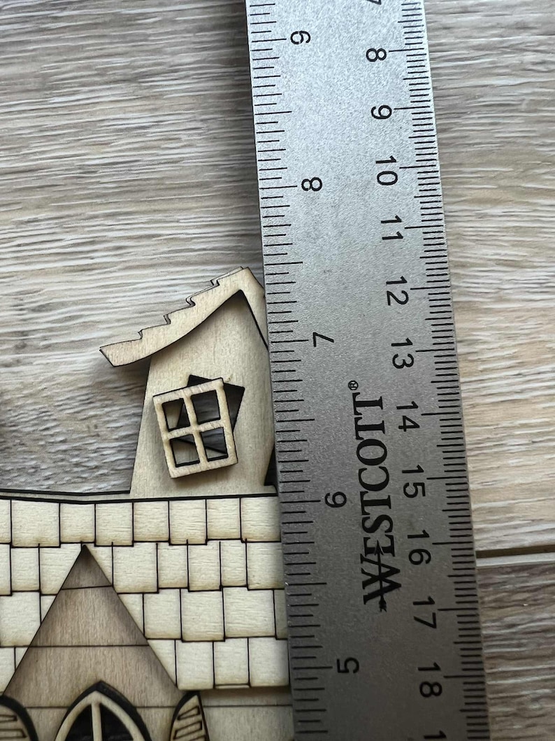 May include: A wooden miniature house with a detailed roof, window, and chimney. A metal ruler is placed next to the house, showing a height of approximately 2.5 inches. The house is made of light-colored wood.
