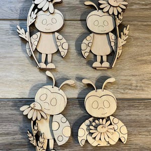 May include: Four wooden ladybug cutouts with daisy and wheat accents. Each ladybug has a smiling face and polka-dot wings. The cutouts are made of light-colored wood and are suitable for crafts or home decor.