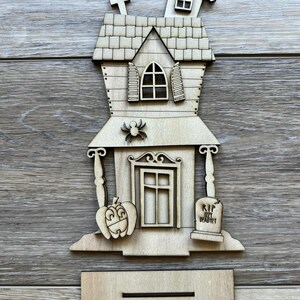 May include: A wooden Halloween-themed haunted house decoration. The house features a pumpkin, spider, and tombstone with the inscription "RIP Izzy DeadYet." Includes a stand for display.