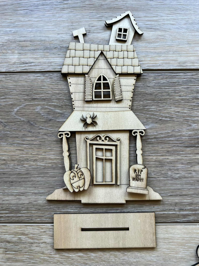 May include: A wooden Halloween-themed haunted house decoration. The house features a pumpkin, spider, and tombstone with the inscription "RIP Izzy DeadYet." Includes a stand for display.