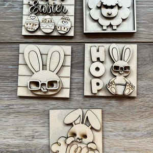 May include: A collection of wooden Easter-themed decorations. The set includes a square sign with "Happy Easter" and decorated eggs, a sheep, two bunnies wearing glasses, a bunny with the word "HOP", and a bunny with curly accents.