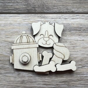May include: Wooden dog-themed cutouts, including a smiling dog face, a fire hydrant, and several bones. The pieces are made of light-colored wood and are arranged on a wooden surface. The dog face has closed eyes.