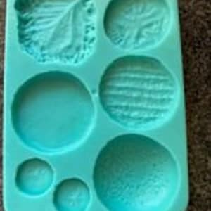 May include: A teal silicone mold with six different shapes: a leaf, two circles, a textured oval, and two smaller circles.