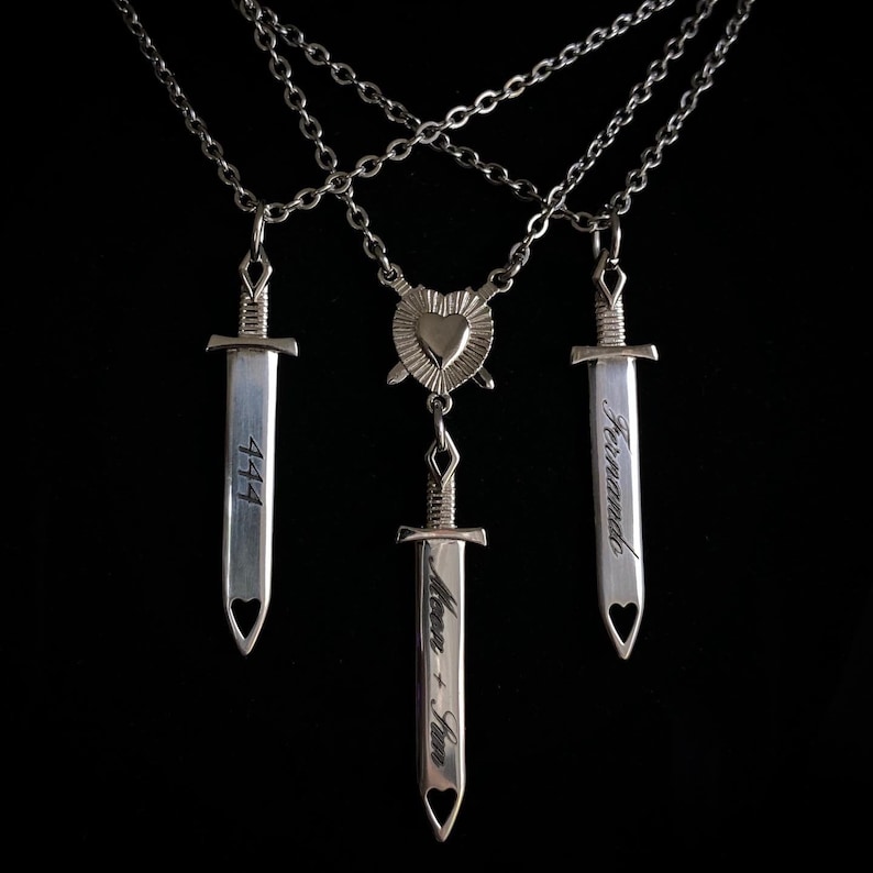 May include: Silver necklaces featuring sword pendants. One pendant is engraved with "444", another with "Fernando", and the third with "Moon + Sun". Each sword has a heart-shaped cutout. A heart-shaped pendant is also included.