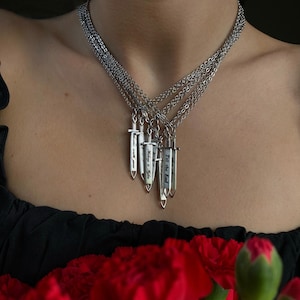 May include: Silver necklaces featuring sword pendants, each inscribed with a different message: "I love you", "Today & Forever", and "Till the End". The necklaces are worn, with red flowers visible in the foreground.