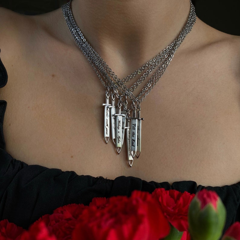 May include: Silver necklaces featuring sword pendants, each inscribed with a different message: "I love you", "Today & Forever", and "Till the End". The necklaces are worn, with red flowers visible in the foreground.