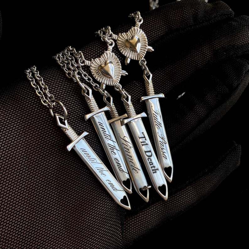 May include: Silver pendant necklaces with sword-shaped charms and engraved phrases. The charms have heart-shaped cutouts and are attached to chain necklaces. Phrases include "until the end", "Finnete", "Til Death", and "Tutto Passa".