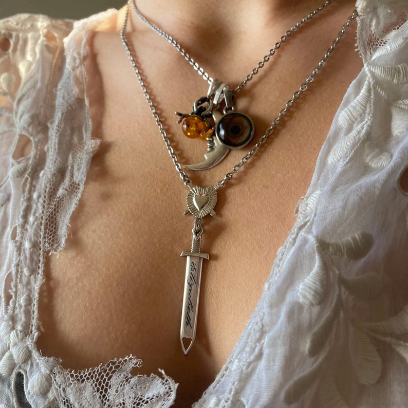 May include: A silver necklace featuring three pendants. The top pendant includes a moon, a brown bead, and a round pendant with an eye design. The middle pendant is a heart with a starburst. The bottom pendant is a sword with the inscription "Elizabeth".