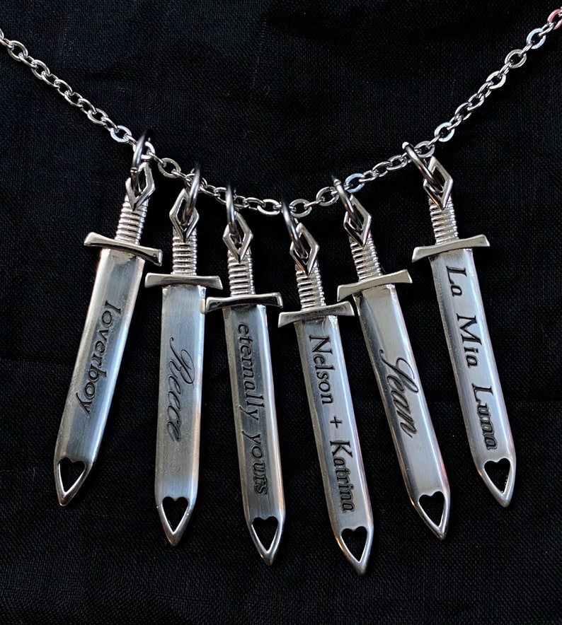 May include: A silver necklace with six sword pendants. Each sword has a heart-shaped cutout and engraved text: "Loverboy", "Reece", "eternally yours", "Nelson + Katrina", "Jean", and "La Mia Luna". The chain is a delicate silver.