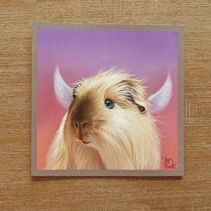 May include: A square greeting card featuring a whimsical illustration of a fluffy, light brown creature with large blue eyes and white horns. The background transitions from pink to purple. The card is set against a wooden surface.