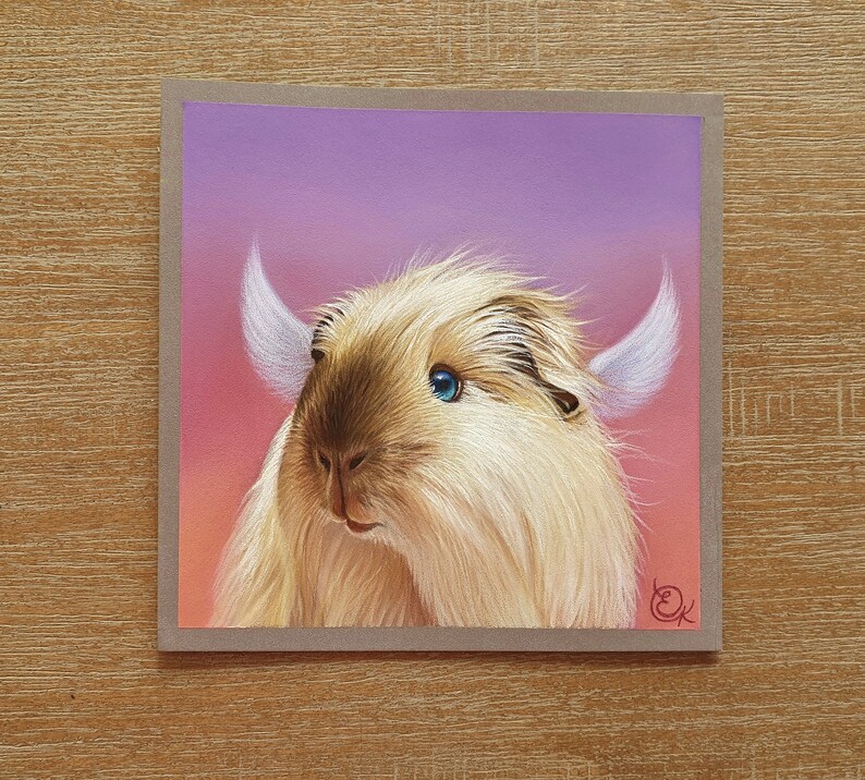 May include: A square greeting card featuring a whimsical illustration of a fluffy, light brown creature with large blue eyes and white horns. The background transitions from pink to purple. The card is set against a wooden surface.