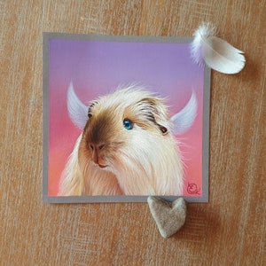 May include: A square artwork featuring a light-colored, furry creature with blue eyes and small horns, set against a pink and purple gradient background. A white feather and a heart-shaped stone are placed near the artwork.