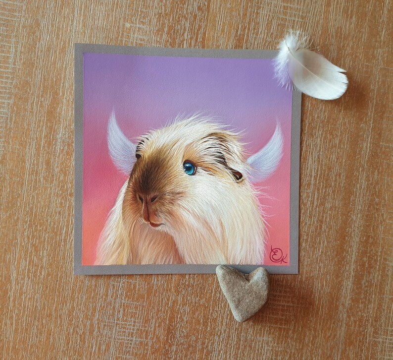 May include: A square artwork featuring a light-colored, furry creature with blue eyes and small horns, set against a pink and purple gradient background. A white feather and a heart-shaped stone are placed near the artwork.